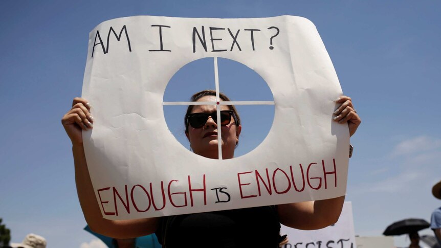 A woman holds a sign with a cut out of a rifle sight over her face