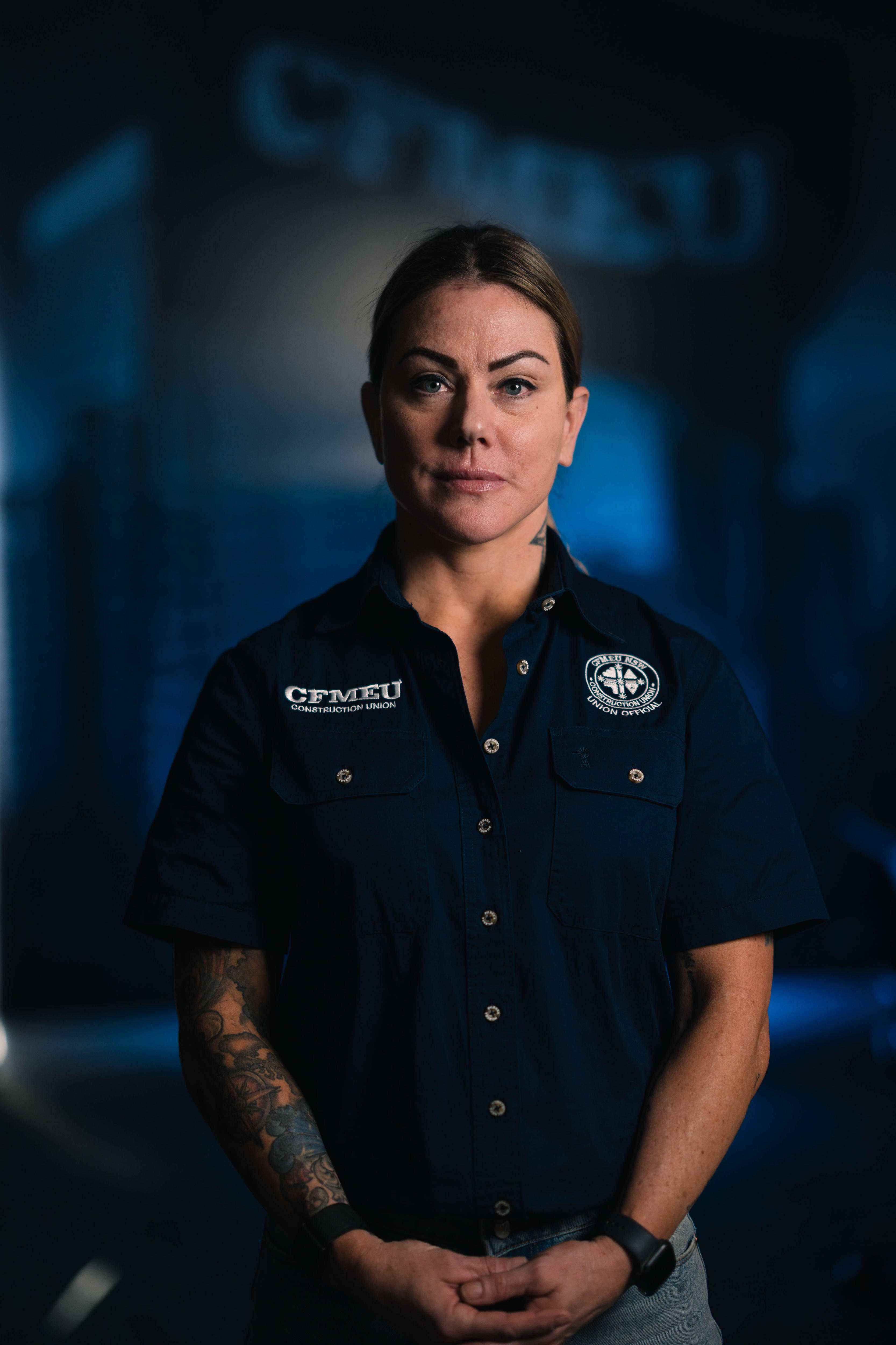 Woman with short hair tucked behind ears stares staunchly at the camera, hands held in front of her, in CFMEU uniform