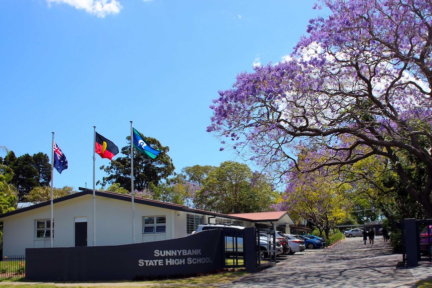 The entrance to Sunnybank State High School with a small white building on the left behind flags next to jacaranda trees