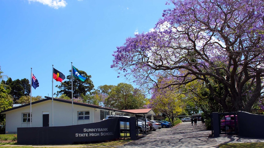 The entrance to Sunnybank State High School with a small white building on the left behind flags next to jacaranda trees