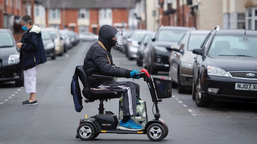 A man with a visor on a mobility scooter in a street with one other lady in it.