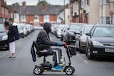 A man with a visor on a mobility scooter in a street with one other lady in it.