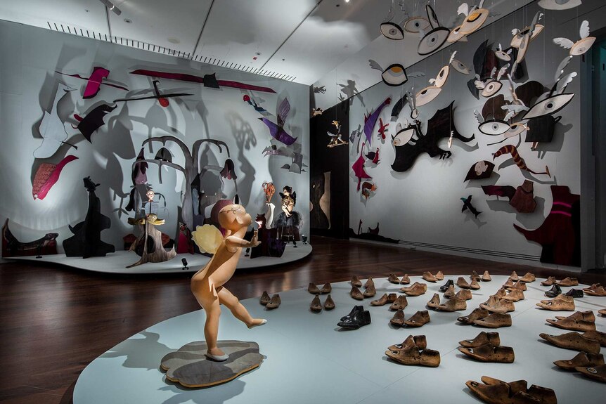 Gallery space with installation in foreground featuring naked child mannequin with wings surrounded by shoes with eyes on them.