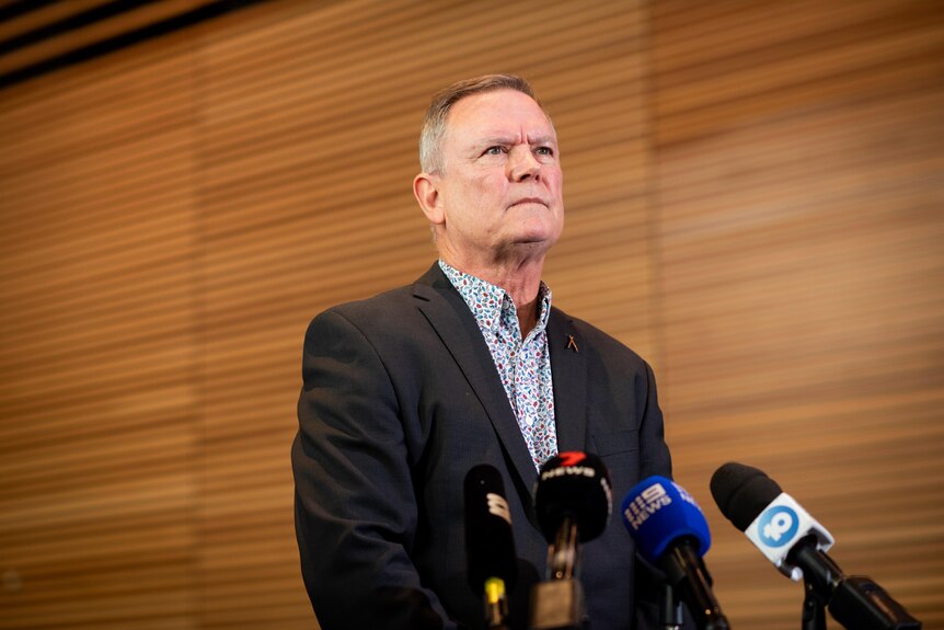 Man in suit and blue patterned shirt addresses media in front of wooden slat wall.