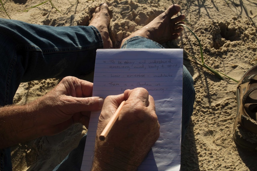 A close up of a man's hand writing on a notebook in pencil while sitting on sand.