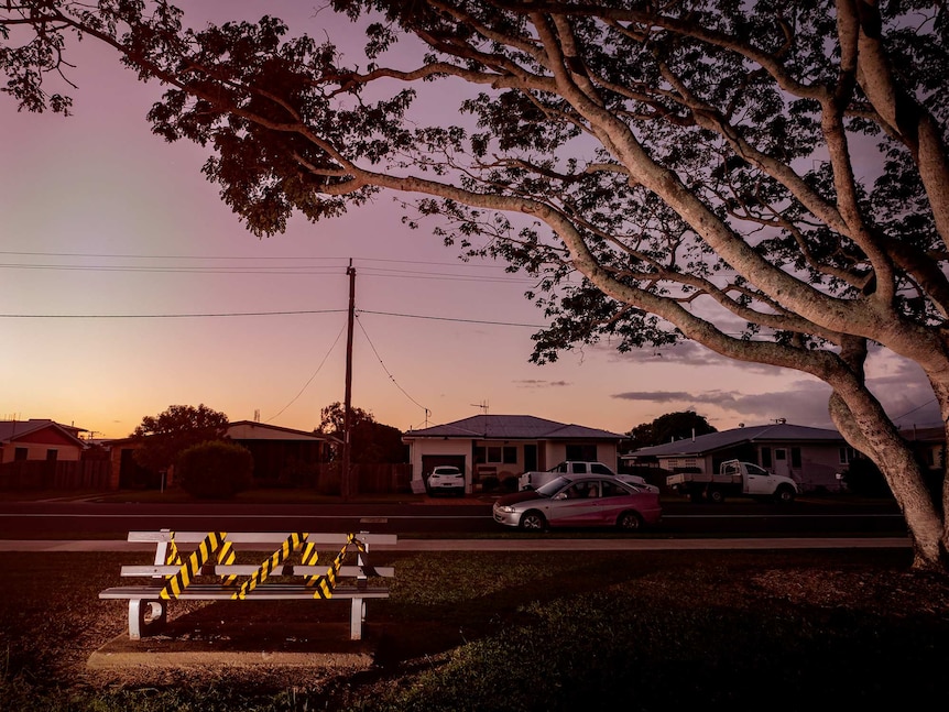 Yellow tape wrapped around a steel bench in a reserve across the road from homes and cars