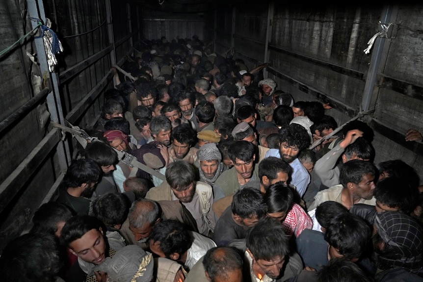 A group of scared-looking men crouch crammed in together in a darkened truck.