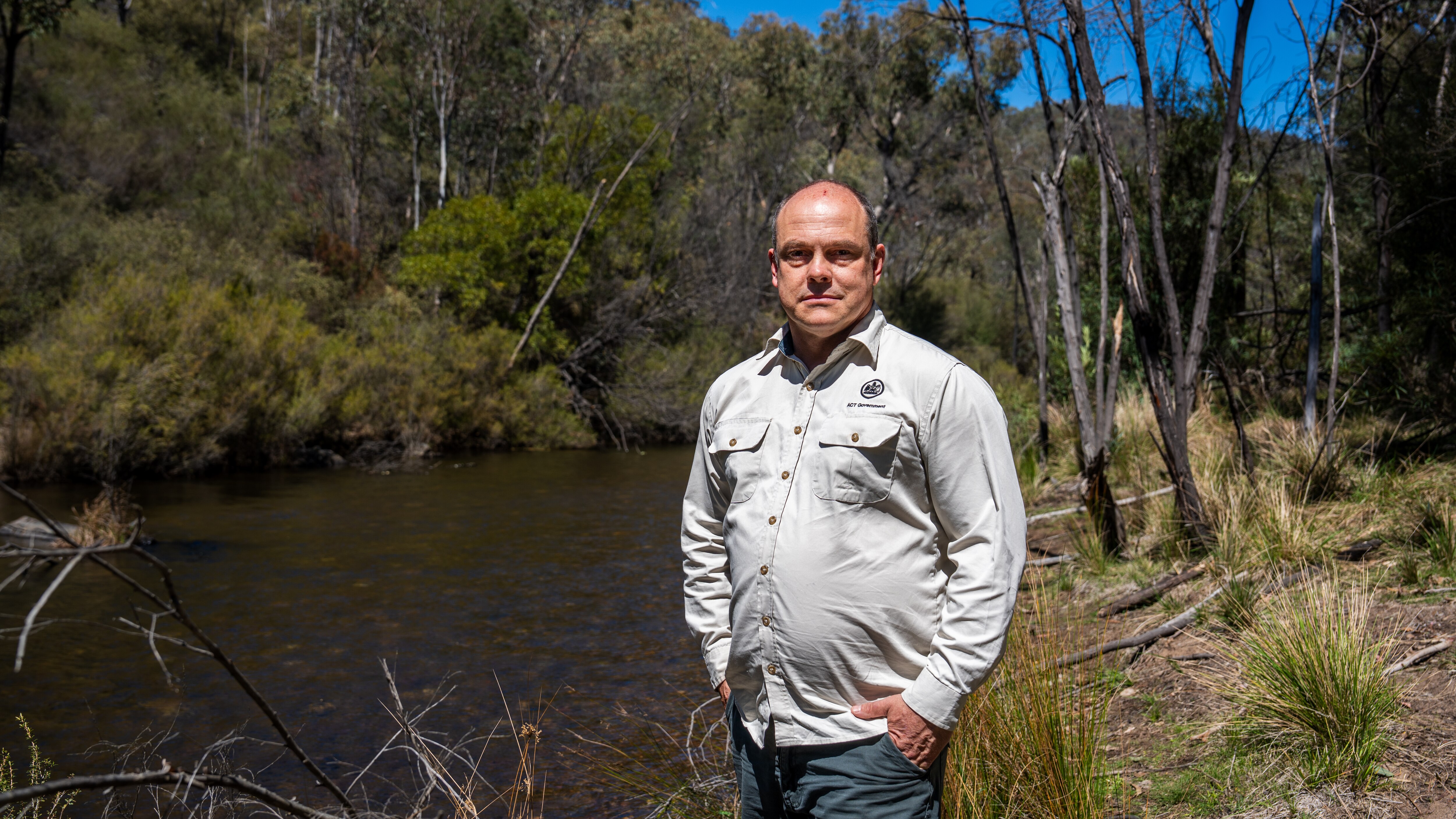 A man wearing a long-sleeved button-down shirt stands on the bank of a slow moving freshwater river in the bush.