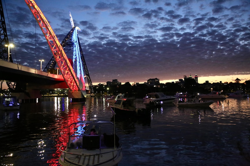 The last of the daylight from a setting sun shows several boats in the Swan River near the Matagarup Bridge lit up blue and red.