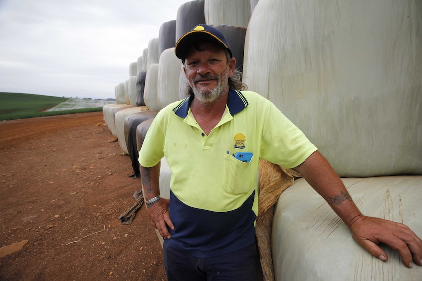 A man in a high vis shirt smiles as he leans on a hay bale in a field.