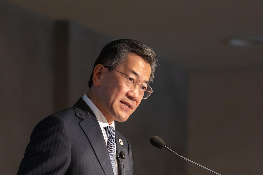 Japanese Ambassador Yamagami Shingo wearing a dark pin-striped suit, glasses, standing at a lectern, speaking seriously.
