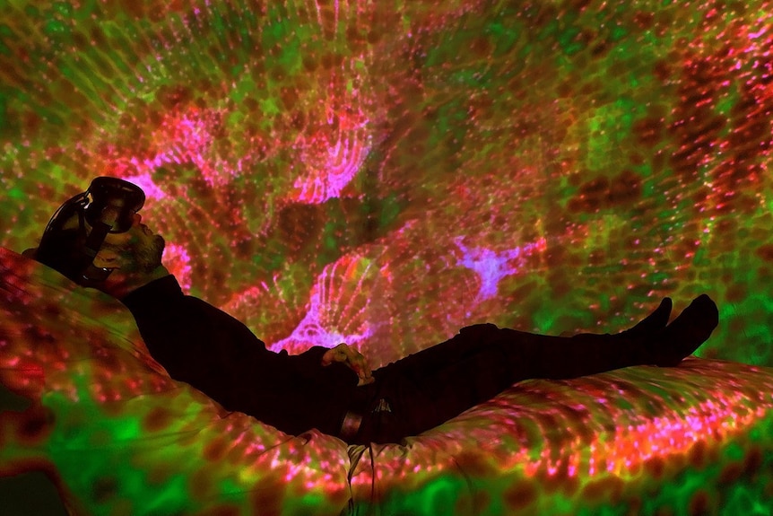Participant lies in interactive bed wearing VR headset, surrounded by kaleidoscopic visuals.