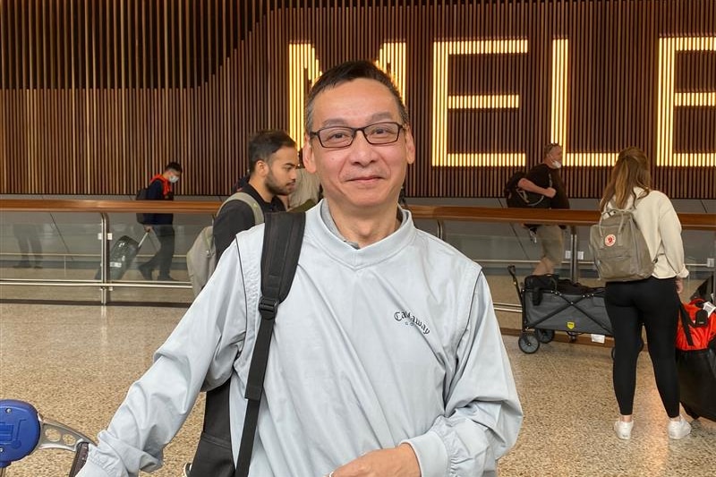 A bespectacled man in a grey top grins as he poses for a photo inside an airport arrivals area.