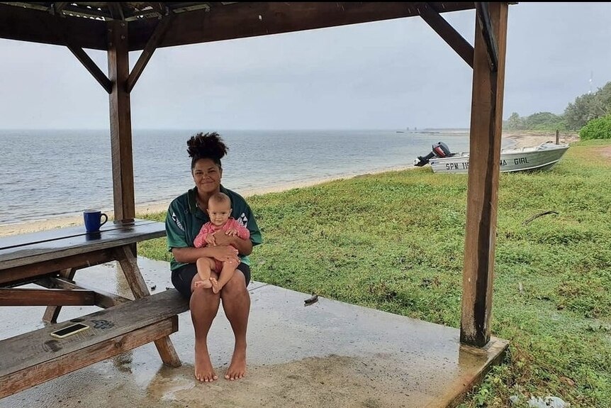 A woman sits on a bench near the ocean with her daughter in her lap