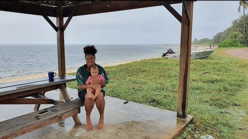 A woman sits on a bench near the ocean with her daughter in her lap