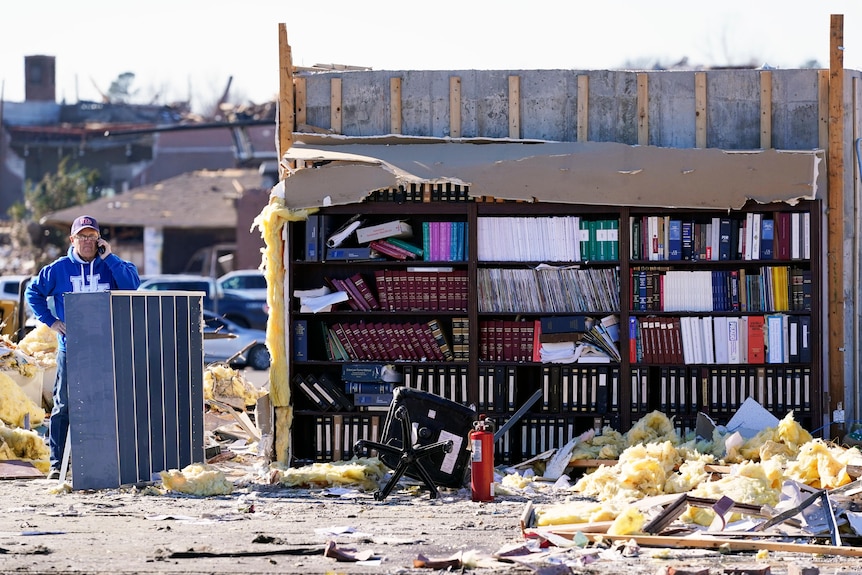 A man speaks on the phone next to a bookshelf standing in debris of a destroyed office