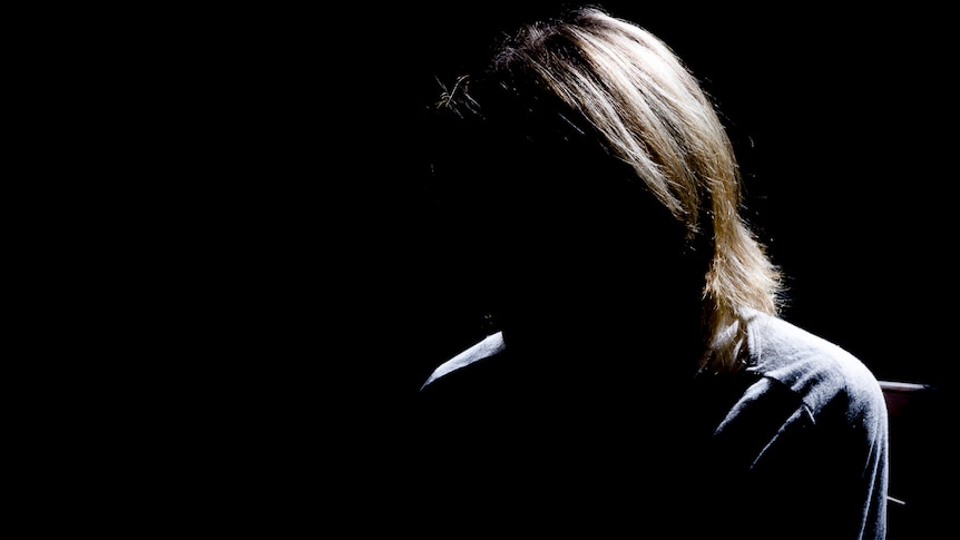 A woman with blonde hair sits in the dark with her face unable to be seen