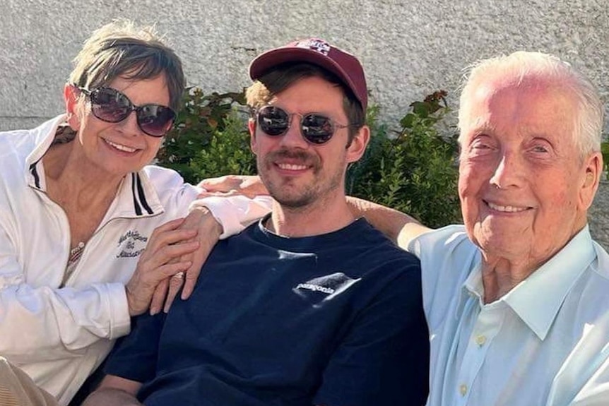 Chris Ross sits between a man and a woman while wearing sunglasses.