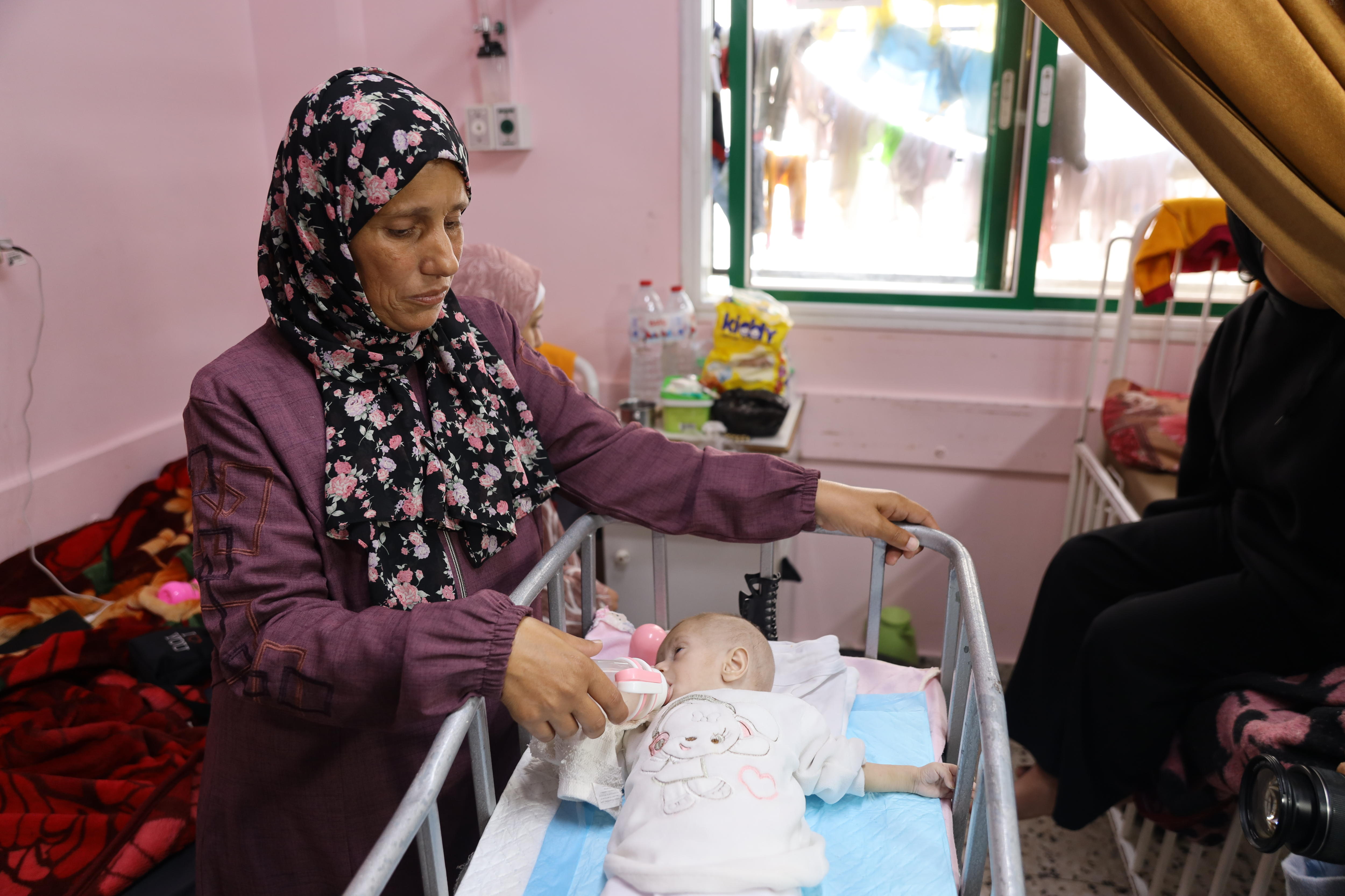 A woman holds a feeding bottle to the mouth of a small baby lying in cot.