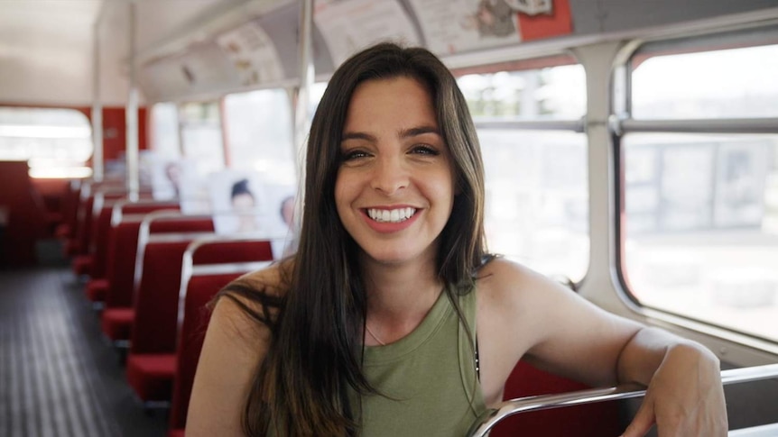 Catalyst presenter Lily Serna sits at the front of a bus, smiling to camera.