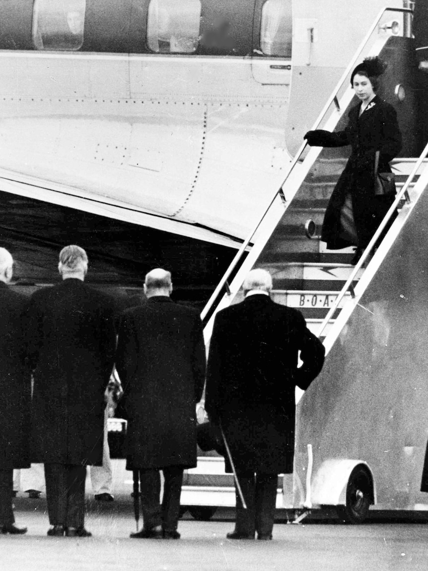 A woman walks down the steps of a plane to a group of men waiting for her