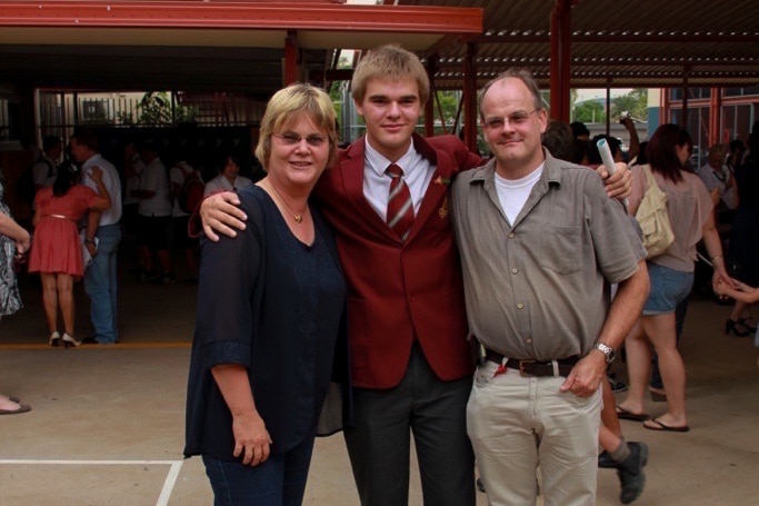 A young man smiles as he poses with his parents at a school graduation.