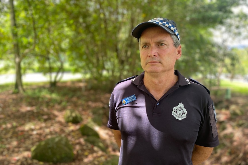 A uniformed policeman with short grey hair stands in a bushy area.