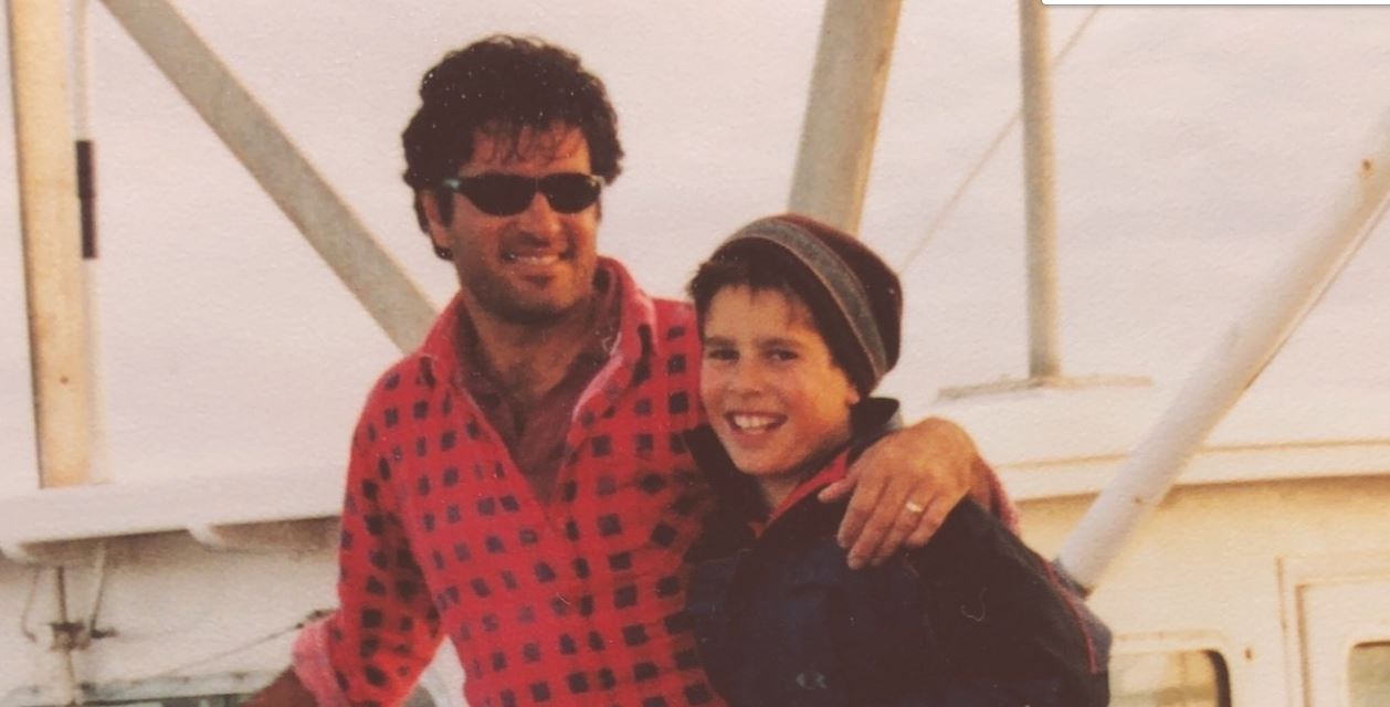 Man smiles with his arm around his son on a fishing boat. The sepia tones on the photo indicate its from the 90s