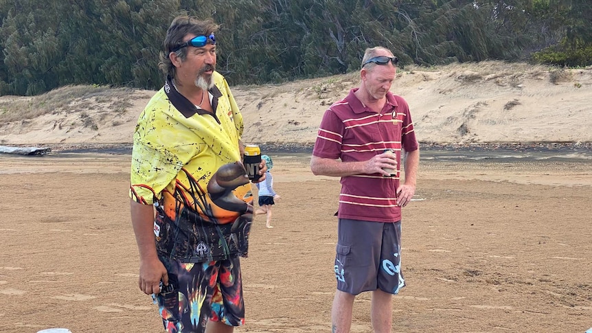 Two men holding cans of beer stand on a beach next