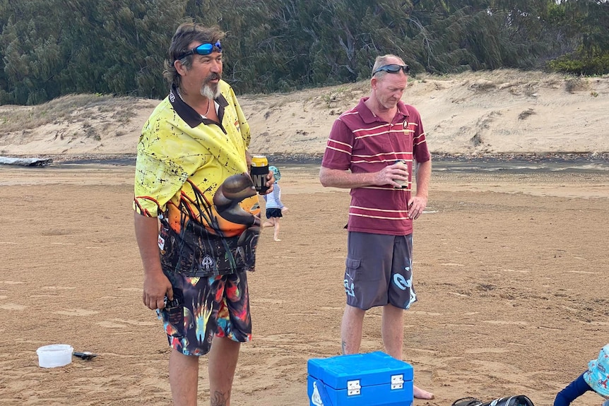 Two men holding cans of beer stand on a beach next