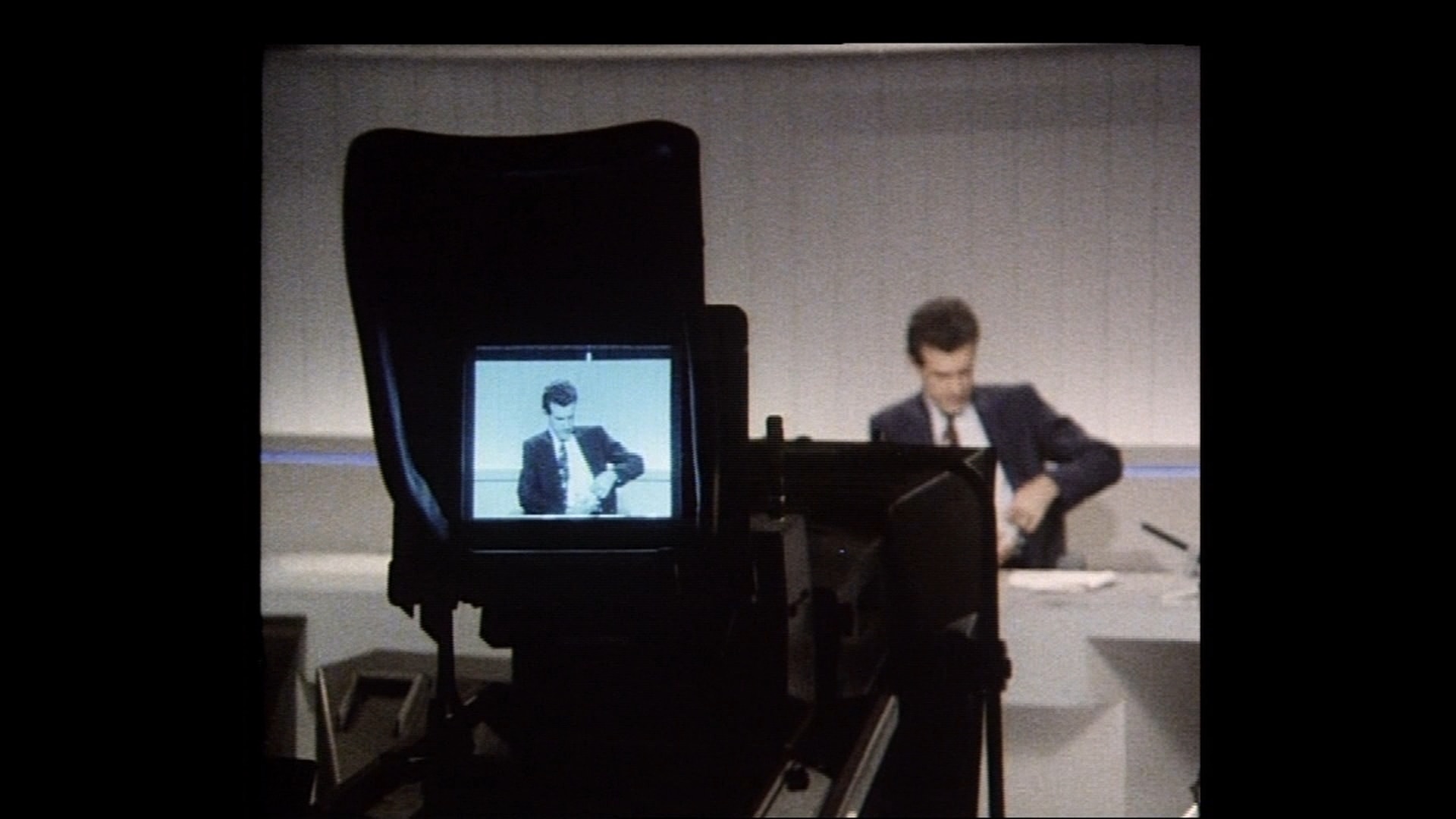 A camera with a screen showing Morecroft in a suit and tie adjusting his shirt at the news desk.