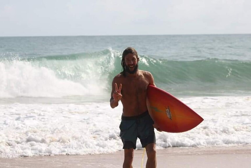 A man with long hair leaves the ocean holding a surfboard, smiling and making a peace sign with his fingers.