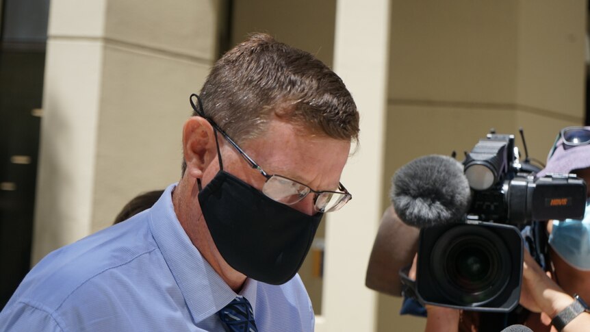 A man with short dark hair and spectacles, wearing a mask, walks away from a court building.