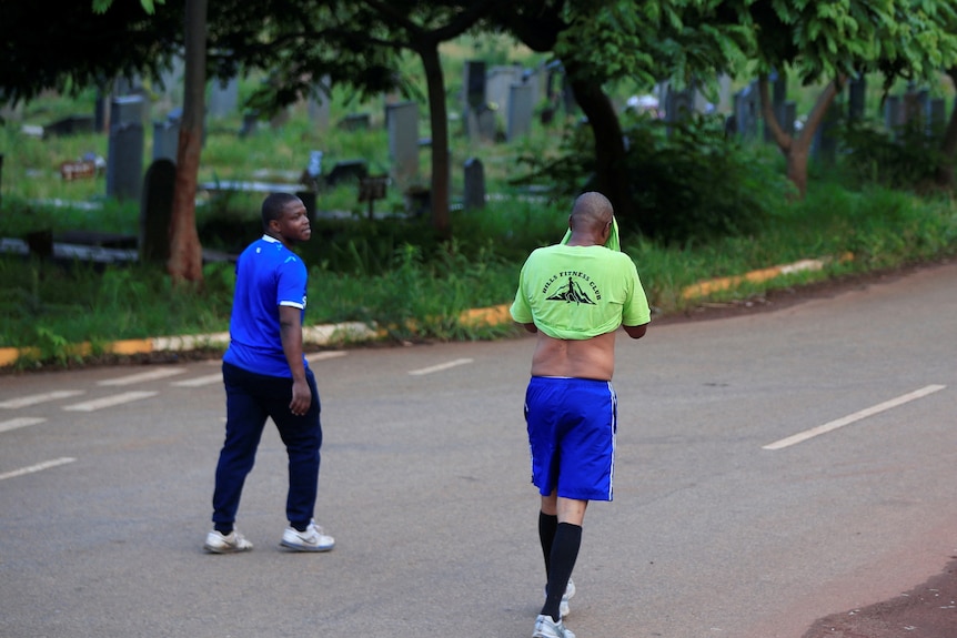 two men in exercise clothing walk along a road next to a cemetery with trees