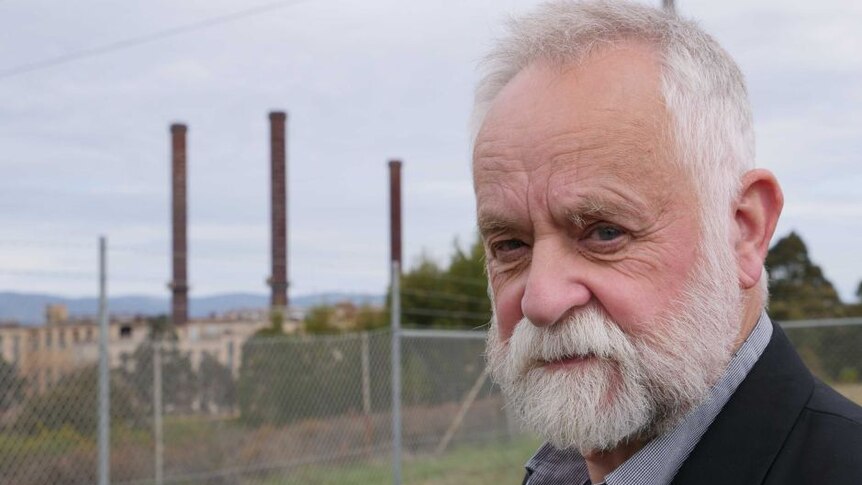 A man with a beard looks at the camera with a power station in the background