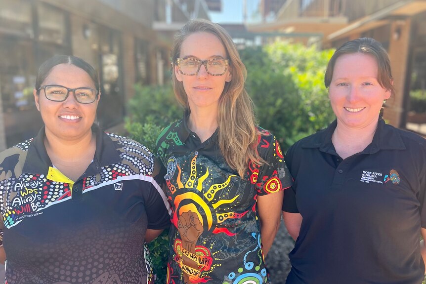 Three women face the camera with their hands behind their backs. They are wearing black shirts with coloured Indigenous prints. 