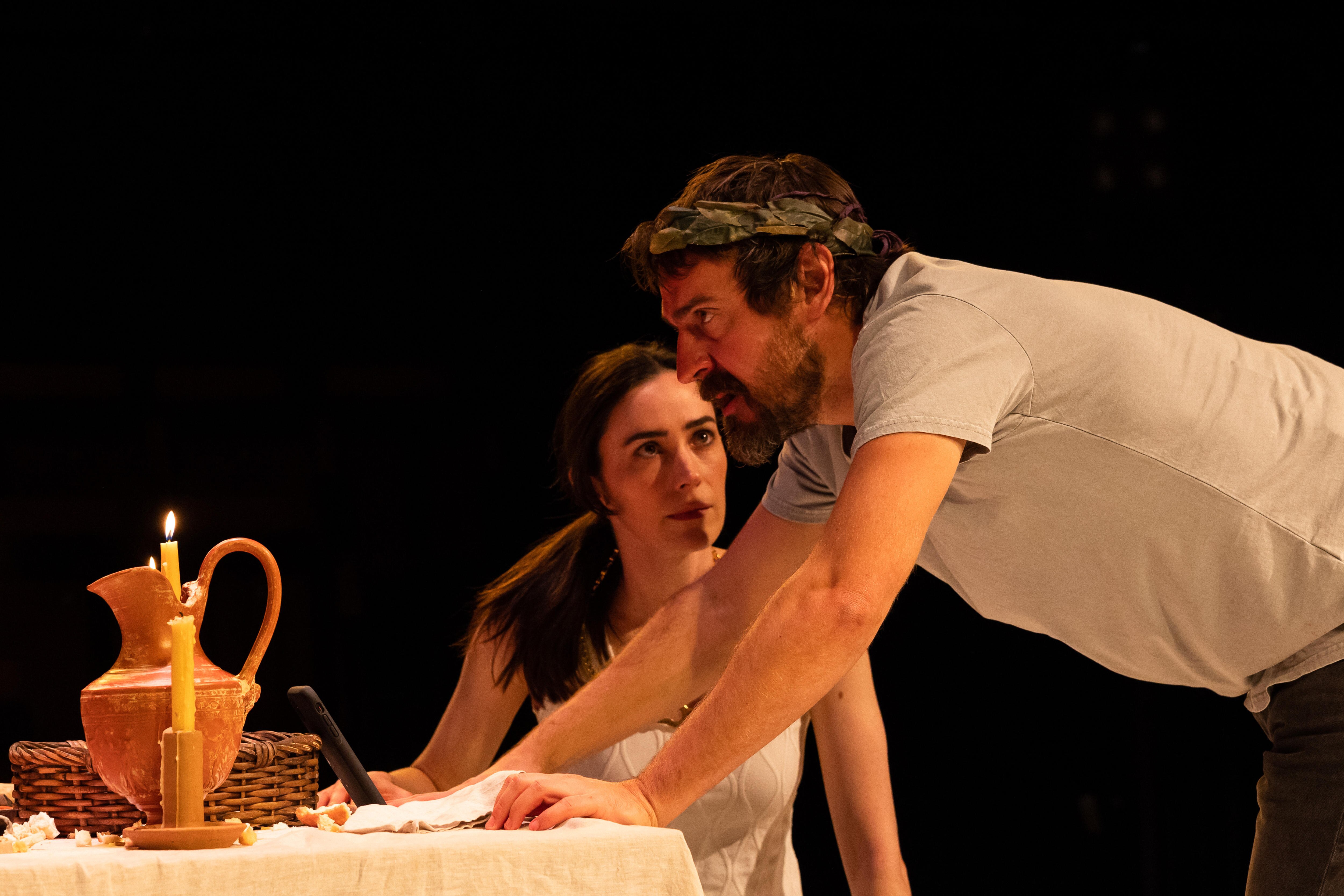 A white woman with dark hair sits at a table and stares intently at a white man wearing a leaf crown.