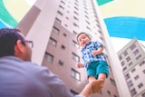 Man lifting up a smiling toddler on his hand, with two apartments in the background behind them.