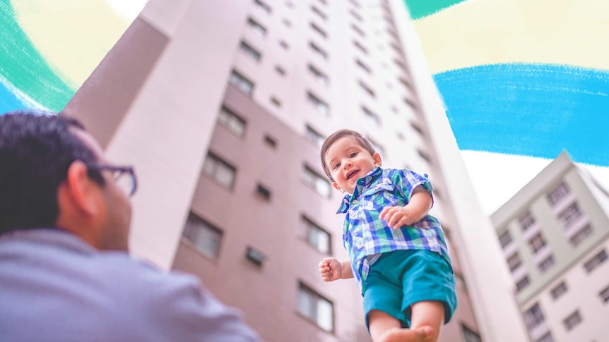 Man lifting up a smiling toddler on his hand, with two apartments in the background behind them.