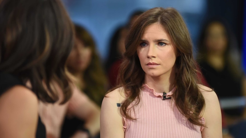 Amanda Knox in a pink top looks at a TV host as she is being interviewed, with audience behind