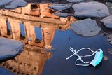 The Colosseum is reflected in a puddle, which has a medical mask sitting in it, on a cobblestone street.
