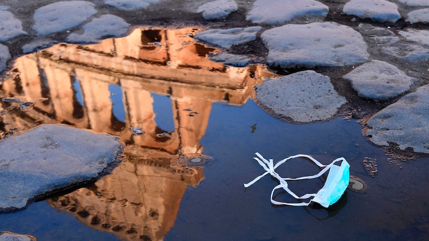 The Colosseum is reflected in a puddle, which has a medical mask sitting in it, on a cobblestone street.