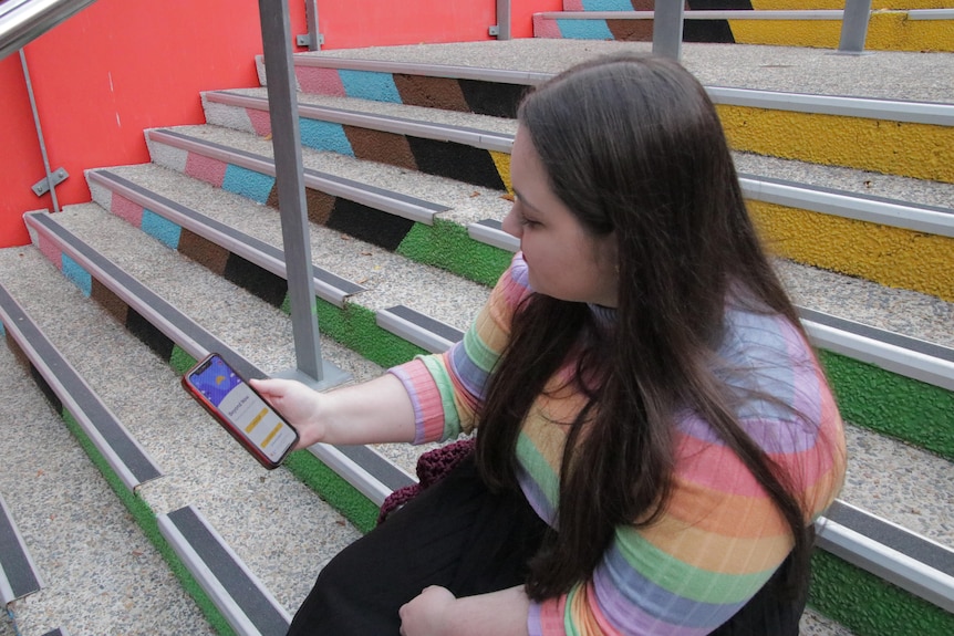 a woman with long brown hair wearing a rainbow coloured top holding a phone.
