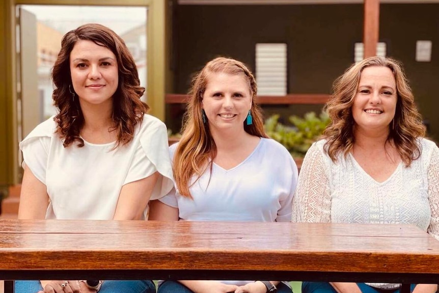 Three women sit at a table in a row wearing white shirts, they're smiling at the camera.