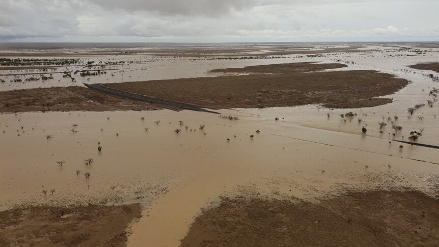 An aerial view of an Outback highway mostly under a brown expanse of floodwater, except for a section of dry land in the middle