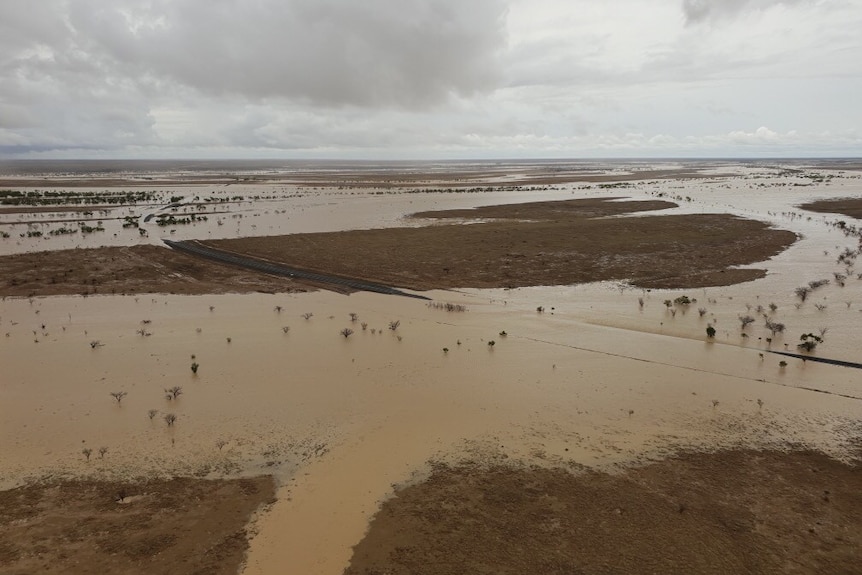 An aerial view of an Outback highway mostly under a brown expanse of floodwater, except for a section of dry land in the middle