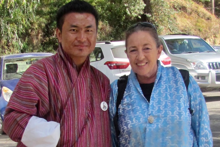An elderly Australian woman poses with a Bhutanese man, both wearing bright Bhutanese clothes.