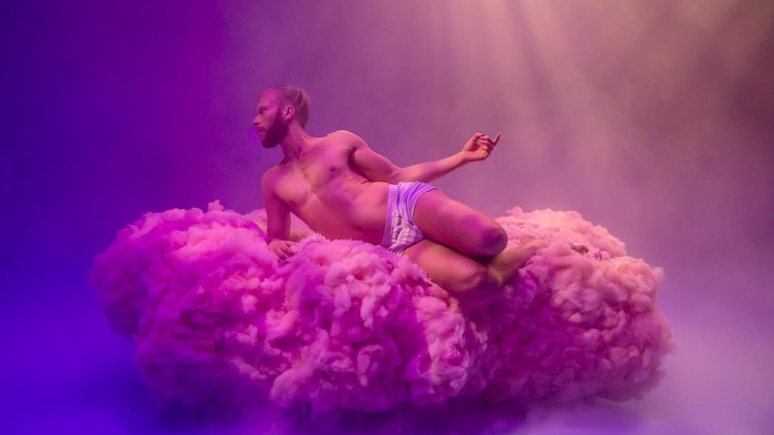 Young man in underwear poses on fluffy pink cloud-like structure, on blue-lit stage.