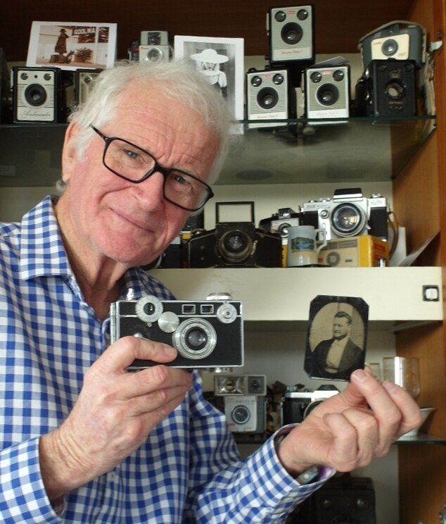 A man with glasses smiles as he holds a film camera and an old photograph.