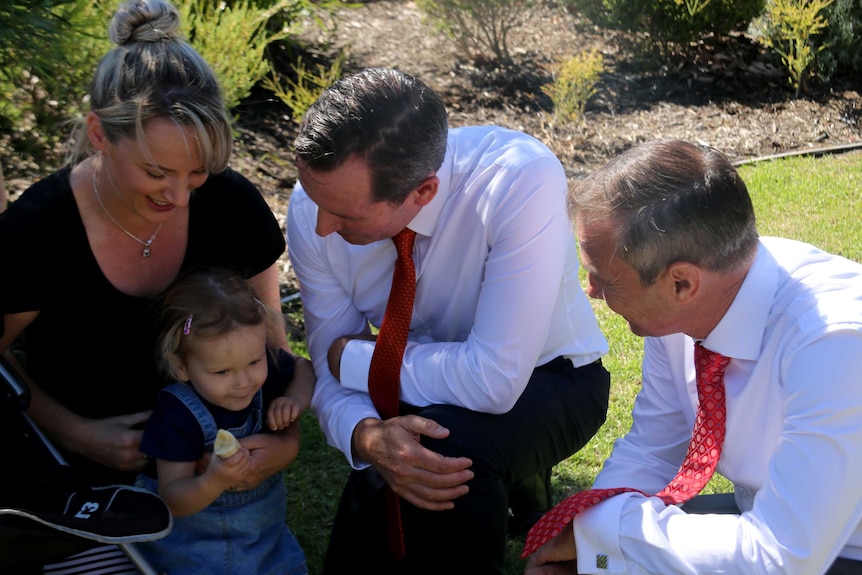 Mark McGowan and Roger Cook speak to a mother and her child crouched on a lawn.